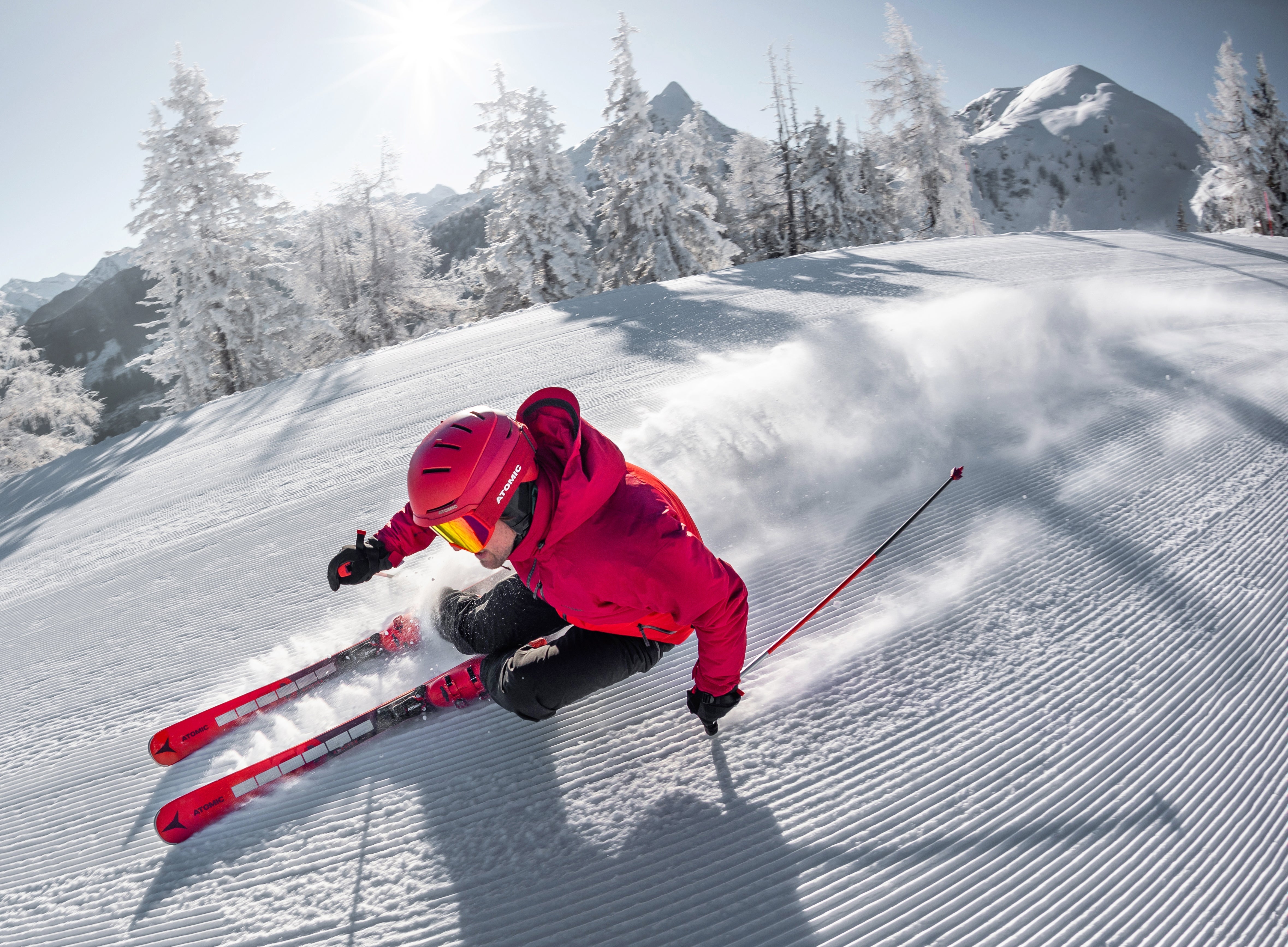 Skier in bright pink jacket carving down groomed slope with snow-covered mountain peaks and frost-covered trees in background