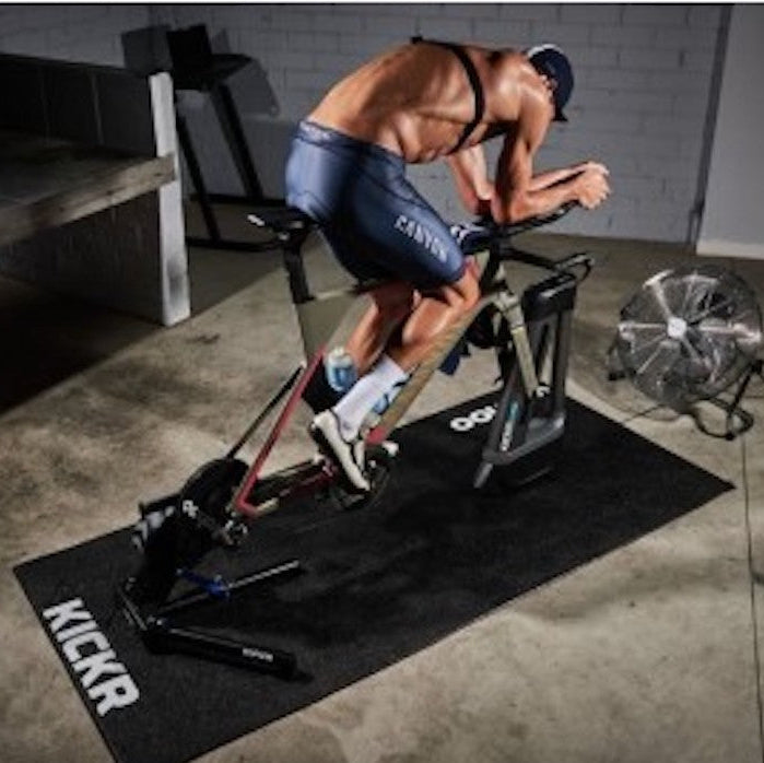 Person using a stationary bike on a KICKR mat in a home gym setting.