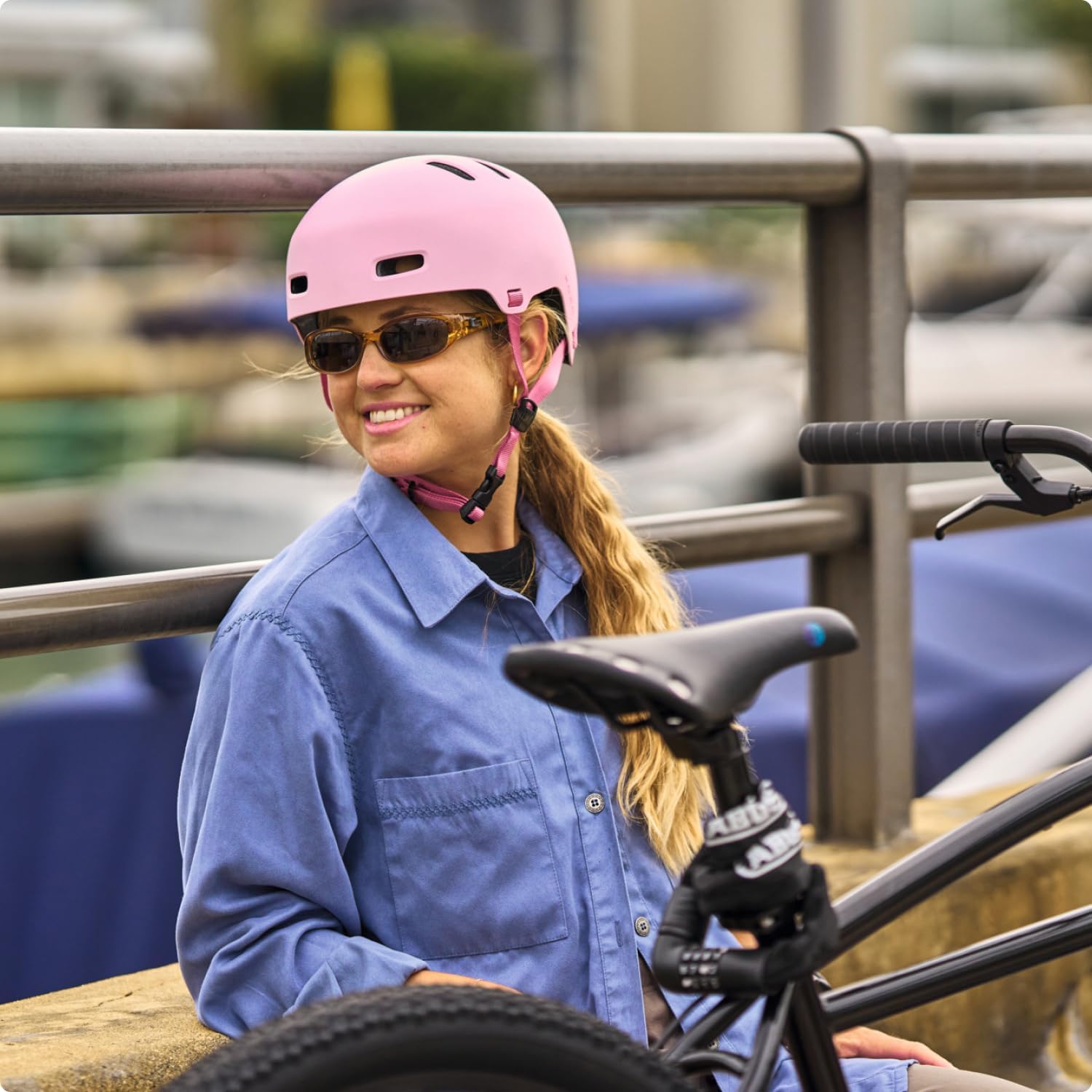 A woman wearing a pink bicycle helmet, sunglasses, and a blue button-up shirt sits next to a black bicycle in an outdoor harbor setting.
