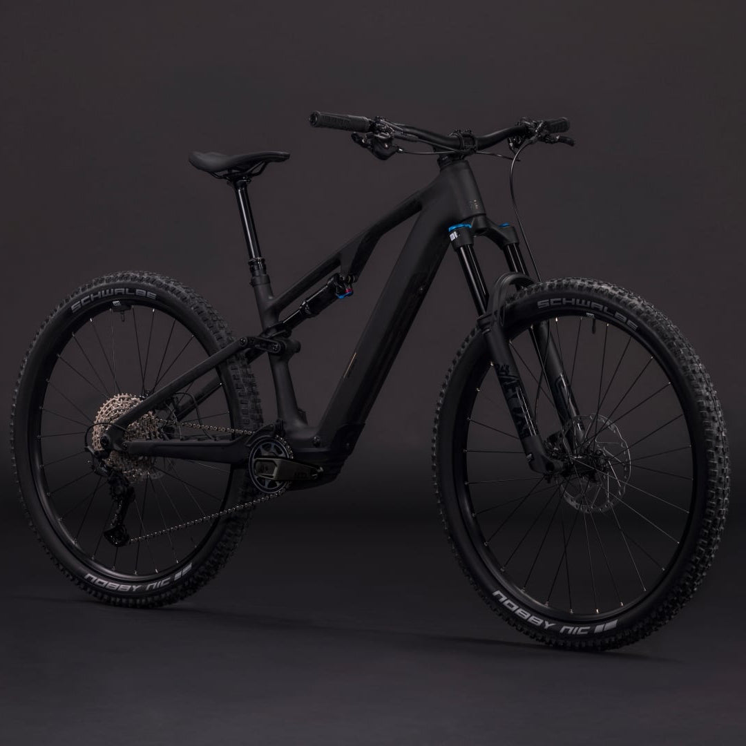 A matte black full-suspension electric mountain bike with knobby tires, a rear rack, and a sleek frame, photographed against a dark gray studio backdrop.