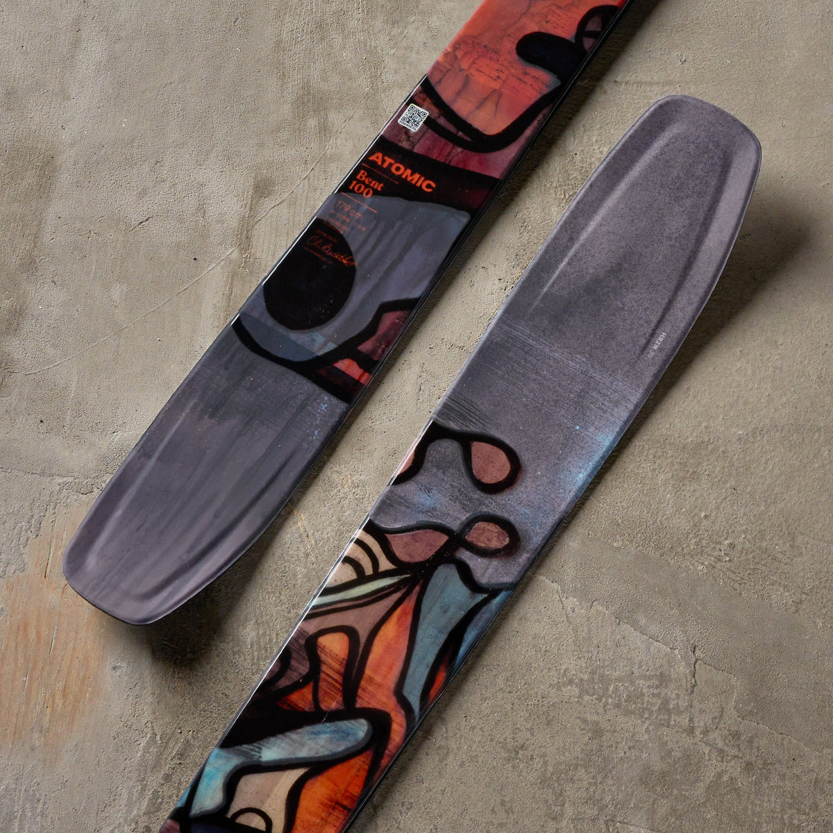 A pair of skis with abstract, colorful graphic patterns in orange, black, and teal on a textured gray background.