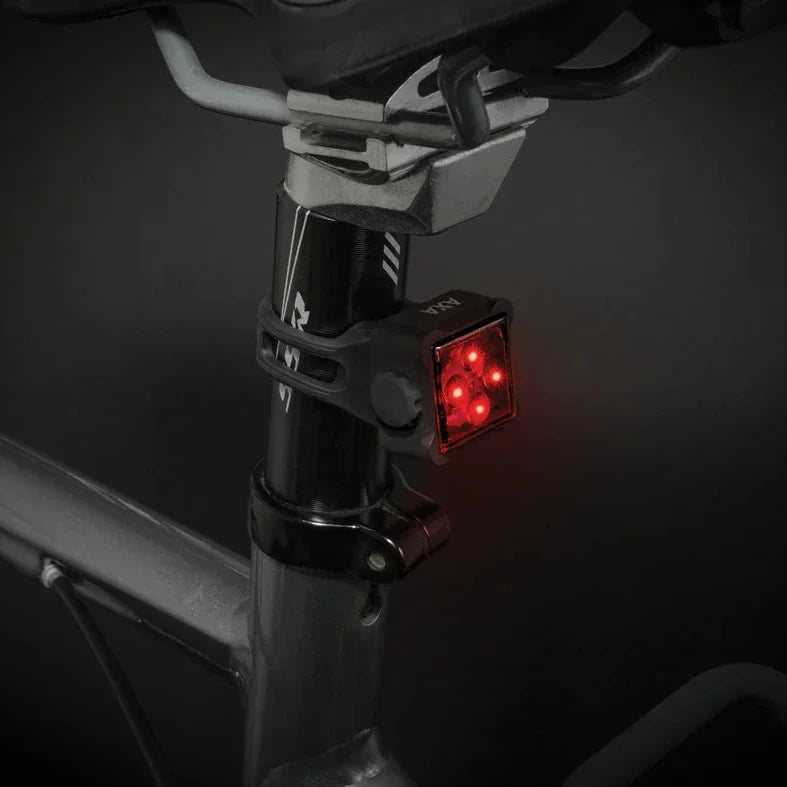 A close-up of a bicycle seat post with a black seat clamp holding a red rectangular rear light, mounted on a dark bicycle frame against a dark background.