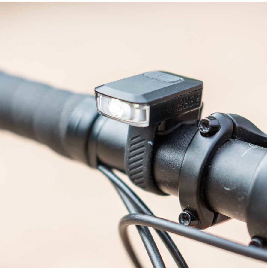 Close-up of a black bicycle handlebar light mounted on a black handlebar with brake cables visible against a blurred beige background.