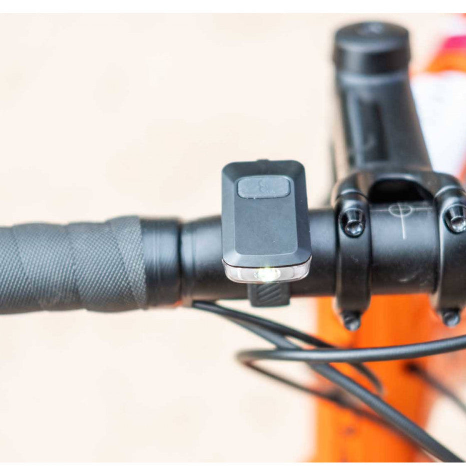 Close-up of a black bicycle handlebar clamp mount holding a small rectangular device, attached to a black handlebar with a textured grip and orange frame visible in the background.