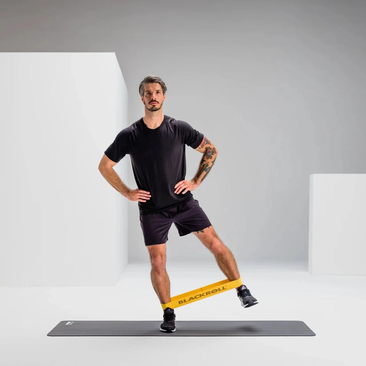 A man in a black athletic shirt and shorts stands on a yoga mat in a studio, using a yellow resistance band around his leg to perform a side lunge. The background is a clean, minimalist gray and white geometric space.