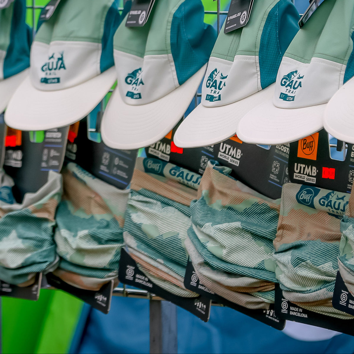 Multiple matching baseball caps hanging on a metal display rack, featuring a light blue and white landscape-style print on the front panels, tan brims, and teal side panels. The caps show the text 'AGA' and 'Menden' on the front, with small black brand tags attached.