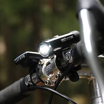 A close-up shot of a black CATEYE bicycle handlebar light mounted on a bike handlebar, with the brand name 'VOLT+COM' also visible on the light body, set against a blurred outdoor background.