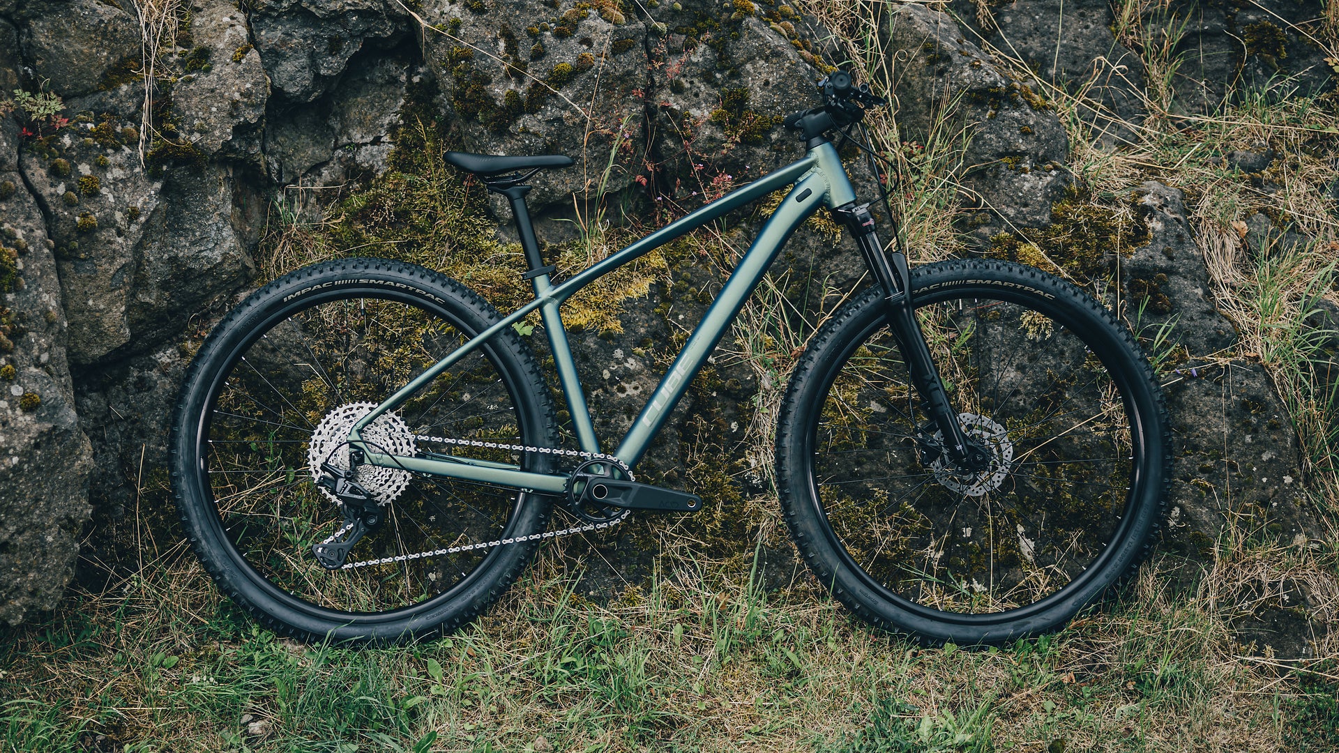 A mountain bike with a matte sage green frame and black components, parked outdoors against a mossy rock wall with grass and dry vegetation.