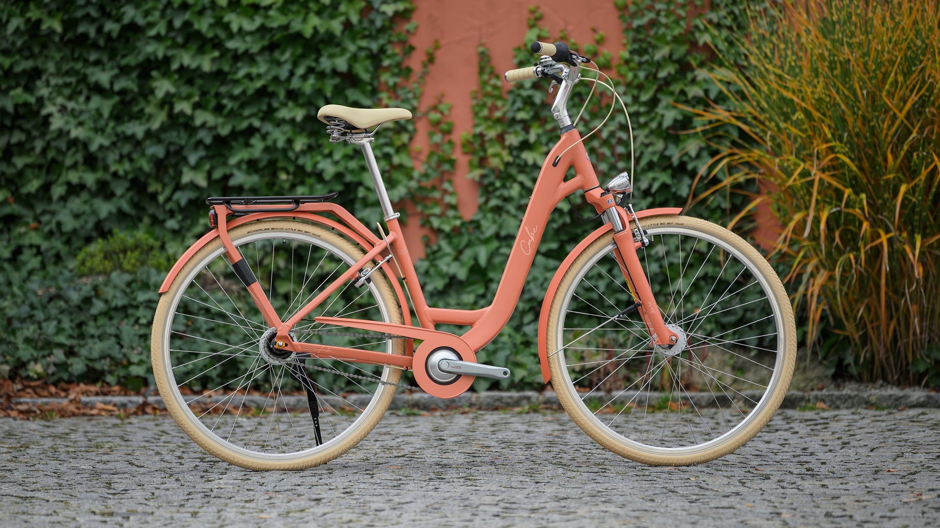 A step-through city bicycle in a muted coral-orange color, featuring cream-colored tires, a beige saddle, and brown grips, parked on a cobblestone surface with a green ivy-covered wall in the background.