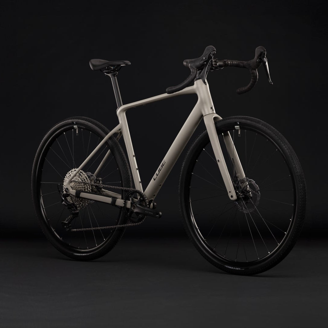 A modern drop-bar bicycle with a matte light gray frame and black wheels, handlebars, and saddle, photographed in a dark studio setting.