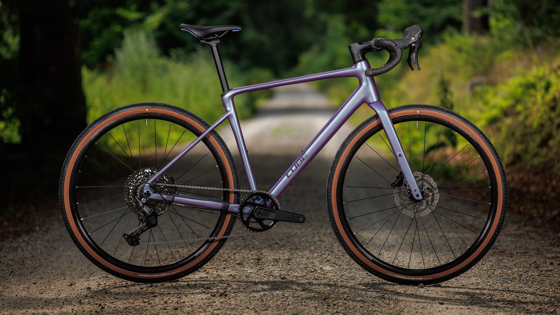 A purple road/gravel bicycle with brown sidewall tires, parked on a gravel path with a blurred green forest background.