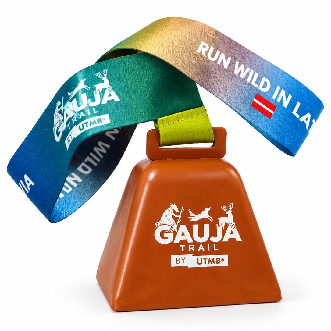 A trail event bell with a brown body and a colorful ribbon lanyard that transitions from blue to green to gold to purple. The bell and ribbon feature the text 'GAUJA TRAIL BY UTMB' and a small Austrian flag.