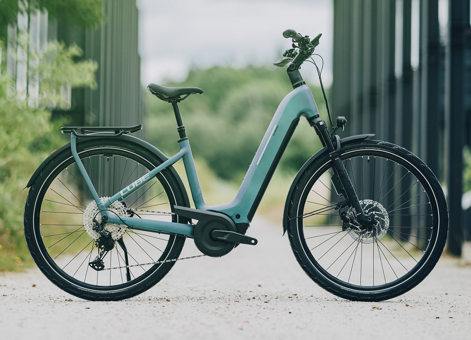 A step-through electric bicycle in a dusty mint green color, parked on a gravel path with a blurred background of green foliage and a dark metal structure.