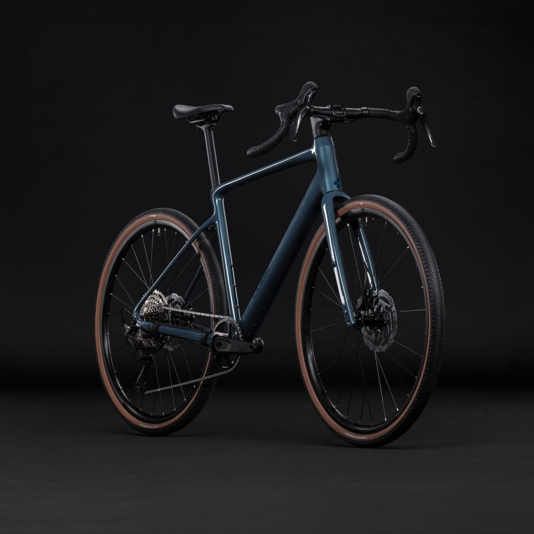 A modern drop-bar bicycle with a matte dark blue-gray frame, black components, and tan sidewall tires, photographed in a dark studio setting.