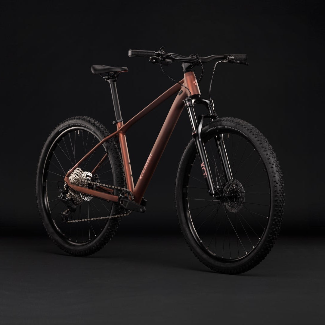 A full-suspension mountain bike with a copper-orange frame and black components, photographed in a studio setting against a dark gray background.