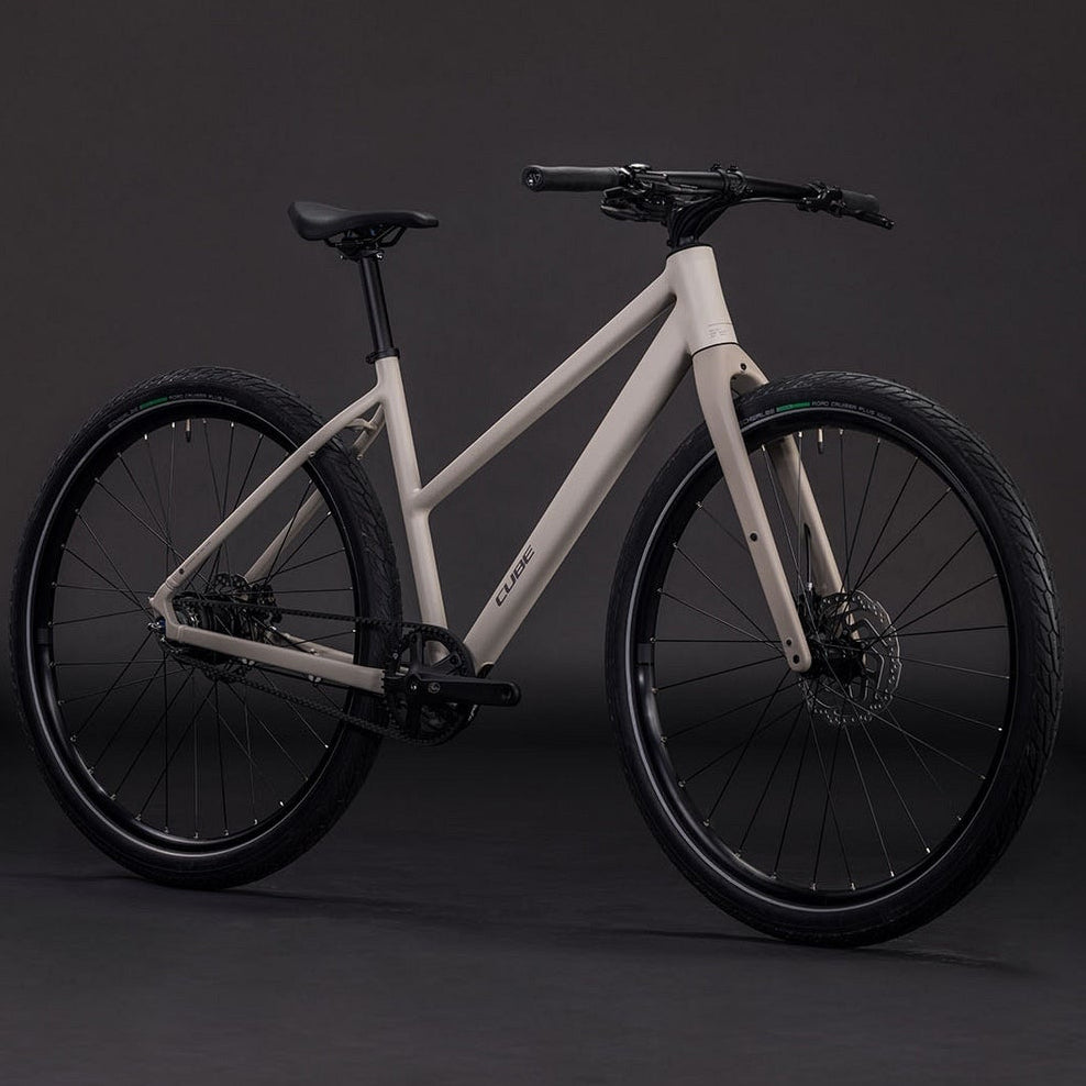 A modern step-through electric bicycle with a matte off-white frame and black components, including tires, wheels, and handlebars, photographed in a studio setting against a dark gray background.