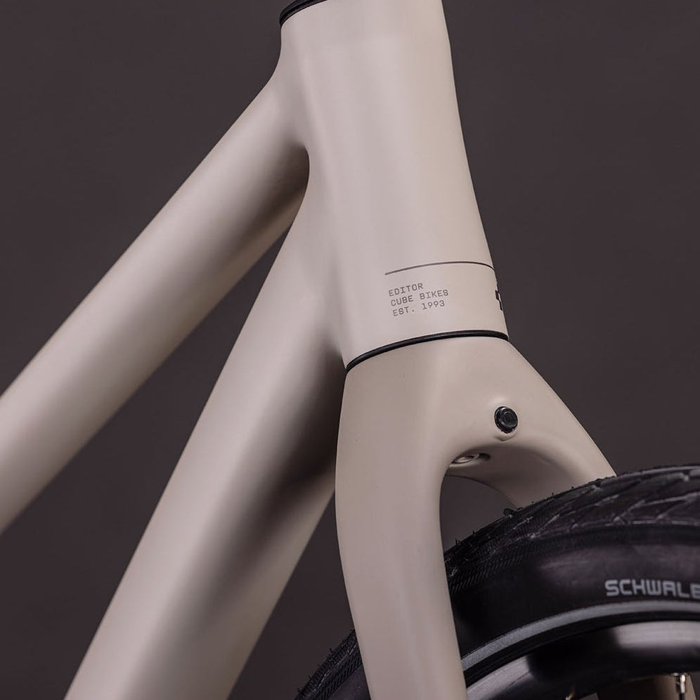 Close-up of a bicycle frame in a matte light beige/taupe color, showing the seat tube junction and part of a black tire against a dark gray background.