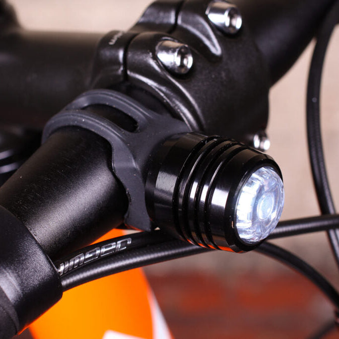 A close-up shot of a black bicycle handlebar light mounted on a black handlebar, with the light turned on and an orange bicycle frame visible in the background.