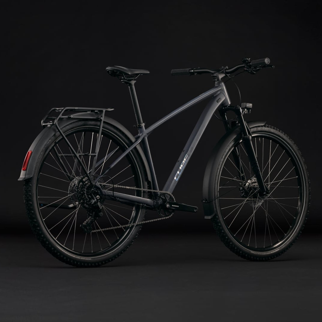 A dark gray electric bicycle with a sleek frame, rear cargo rack, and fenders, photographed in a side profile against a dark studio background.